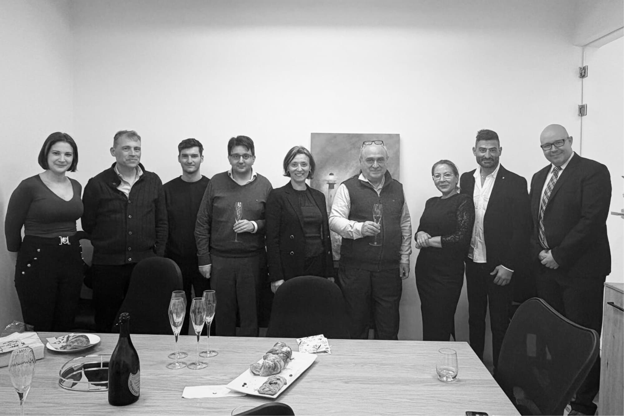 Black-and-white photo of nine people gathered in an office, each holding a champagne glass, with pastries and flutes on the table in front of them.