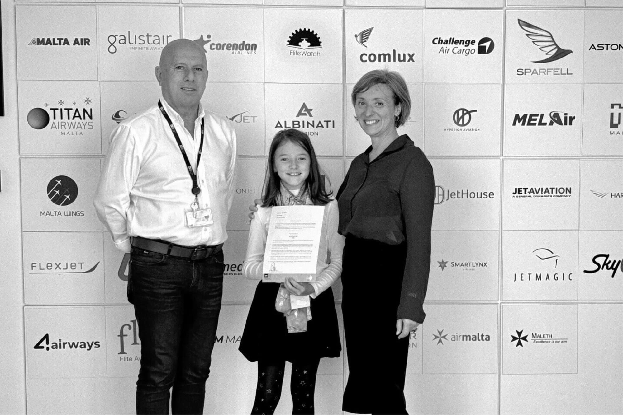 Black-and-white photo of two adults and a child standing in front of a backdrop covered with aviation logos. The child holds a document while all three smile at the camera.