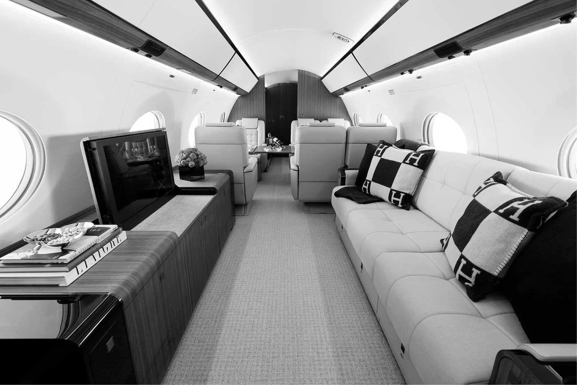 Luxurious private jet interior with plush cream-colored seating, black and white Hermes pillows, a built-in TV, wooden cabinetry, and elegant lighting.